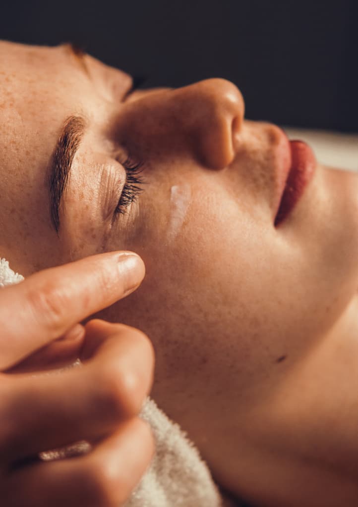 Visage d'une femme avec les yeux fermés se faisant appliqué de la crème sur les pommettes par la spa praticienne