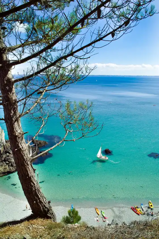 Plage de galets en Bretagne avec eau turquoise, voilier et kayaks — évasion bien-être signée Namaspa.fr